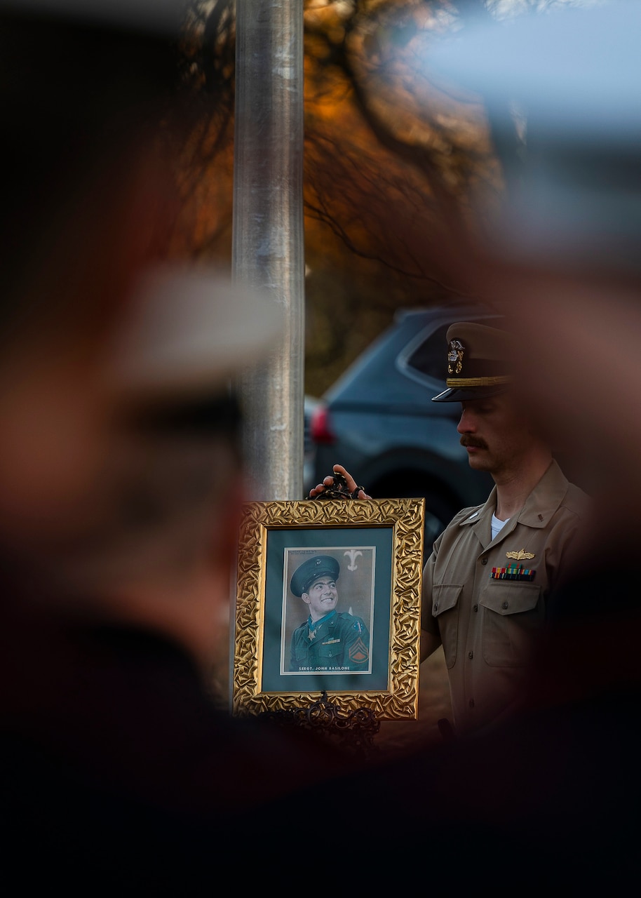A sailor is seen through the saluting arm of another, holding a framed image of another sailor.