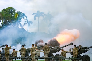 Smoke fills the air as some soldiers fire cannons as others salute while standing in formation.