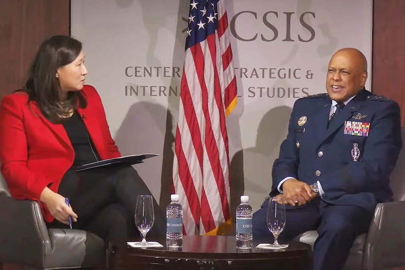 A woman and a man speak while sitting at a table on a stage.