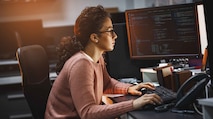 A curly haired woman with glasses sitting at a desk, typing on the computer and looking at a computer screen that is displaying data