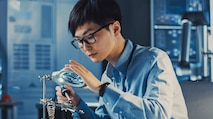 An Asian man sitting in a lab at a desk using a microscope to work on a small piece of machinery