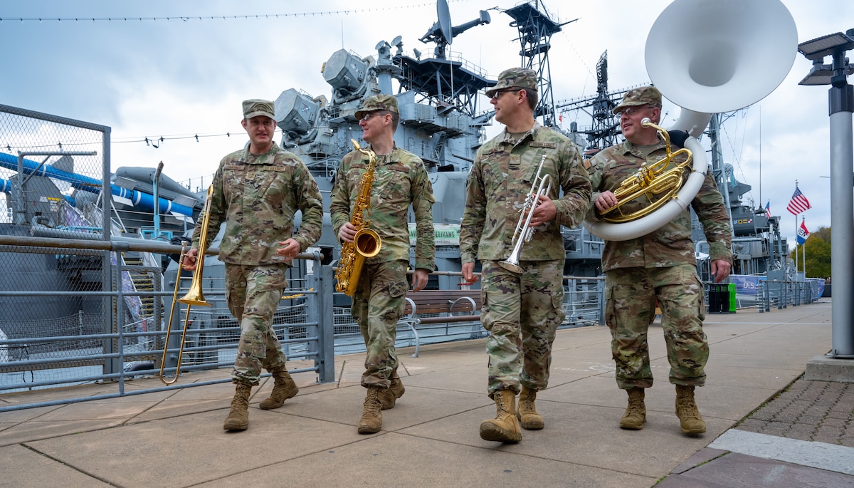 Four uniformed Air Force band members walk in front of a naval ship carrying instruments.