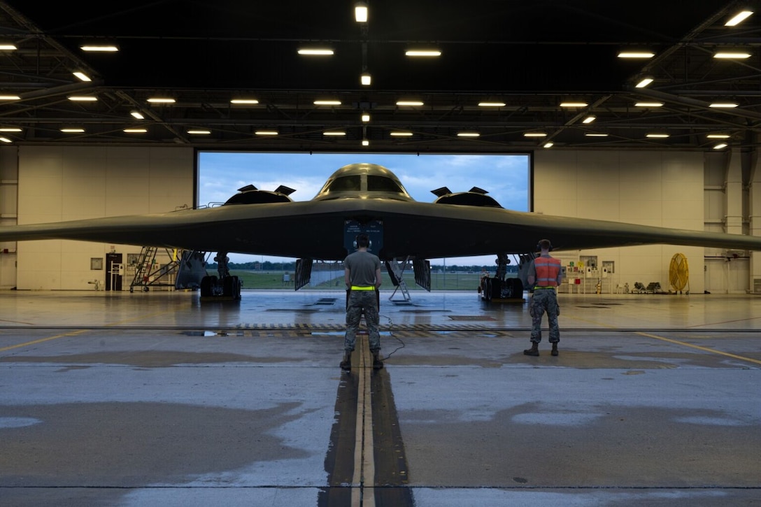 Two men in military uniforms attend to a large stealth aircraft in a hanger.