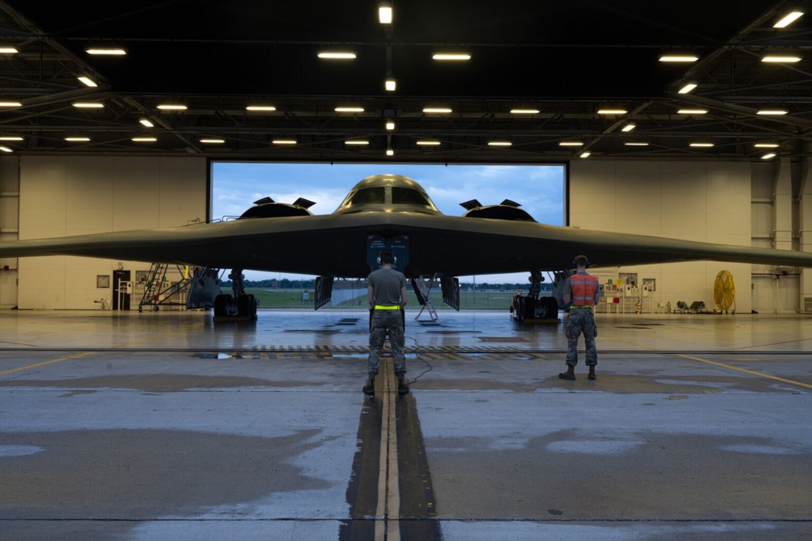 Two men in military uniforms attend to a large stealth aircraft in a hanger.