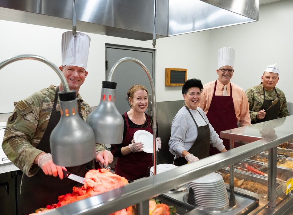 A group of five people stand behind a food service line. Two of them in chefs' hats.