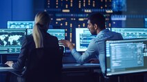 A blonde-haired woman and an Indian man are facing their computer screens in a dark lab and talking to one another about the data they are seeing