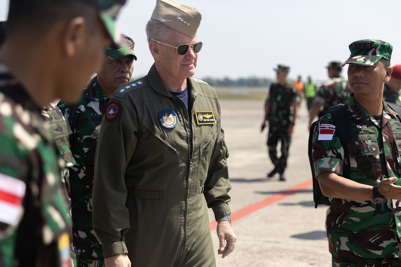 A navy admiral walks among partner service members on tarmac outside.