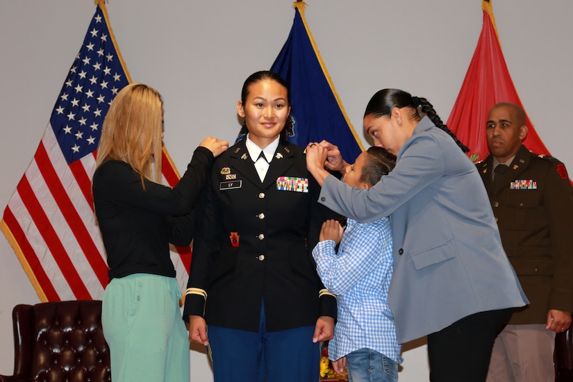 A soldier standing in front of flags receives pins on each shoulder from family members while a service member stands in the background.