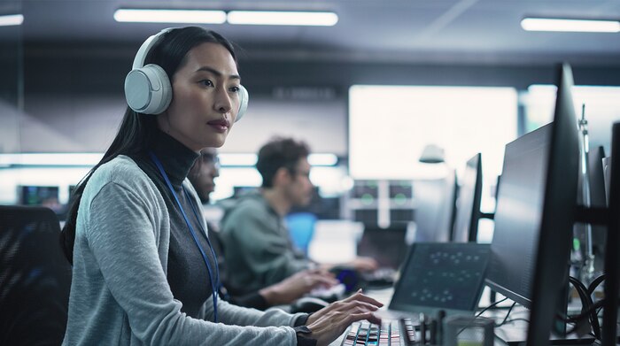 An Asian woman wearing large white headphones, looking at multiple computer screens in front of her
