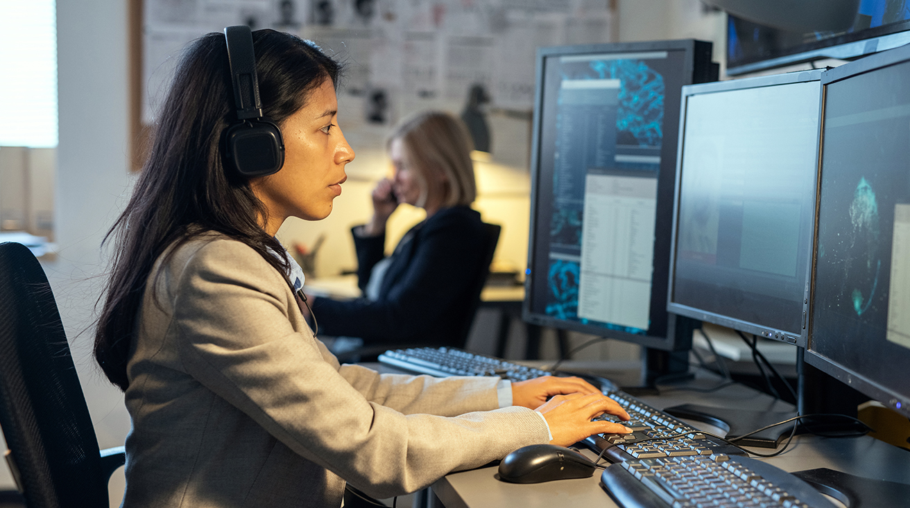 A woman sitting at a desk with multiple computer screens, typing while listening to transcripts with a coworker on the phone in the background.