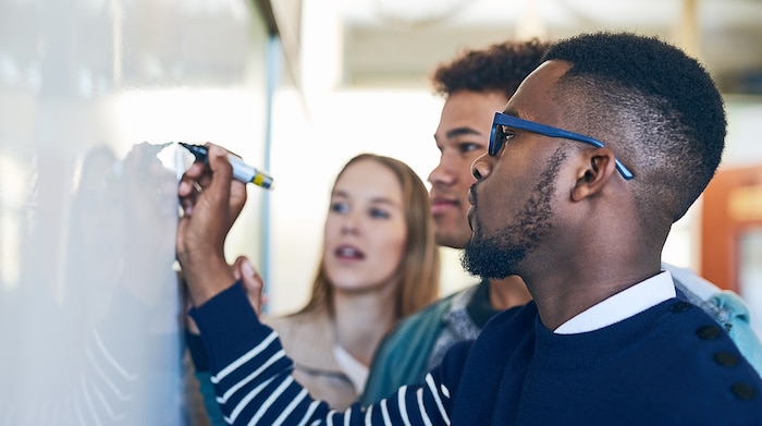 A black high school male with glasses writing on a whiteboard and standing next to two other students who are focused on what he is writing