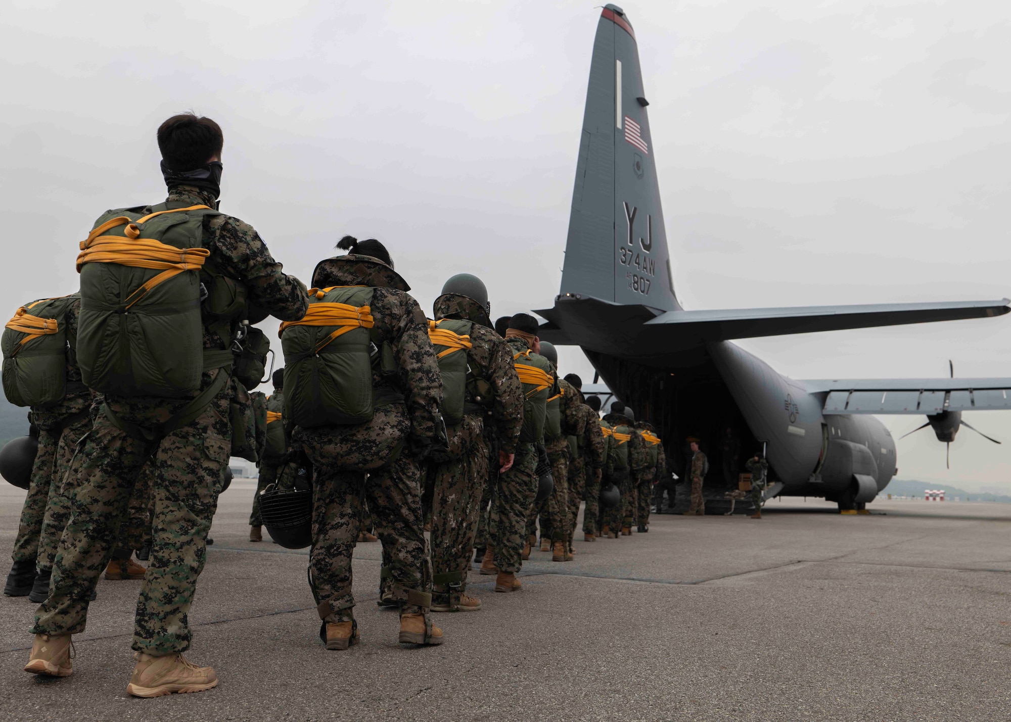 A line of soldiers in uniform and wearing parachute rigging stand on a flightline behind a C-130J aircraft with it's cargo ramp down.