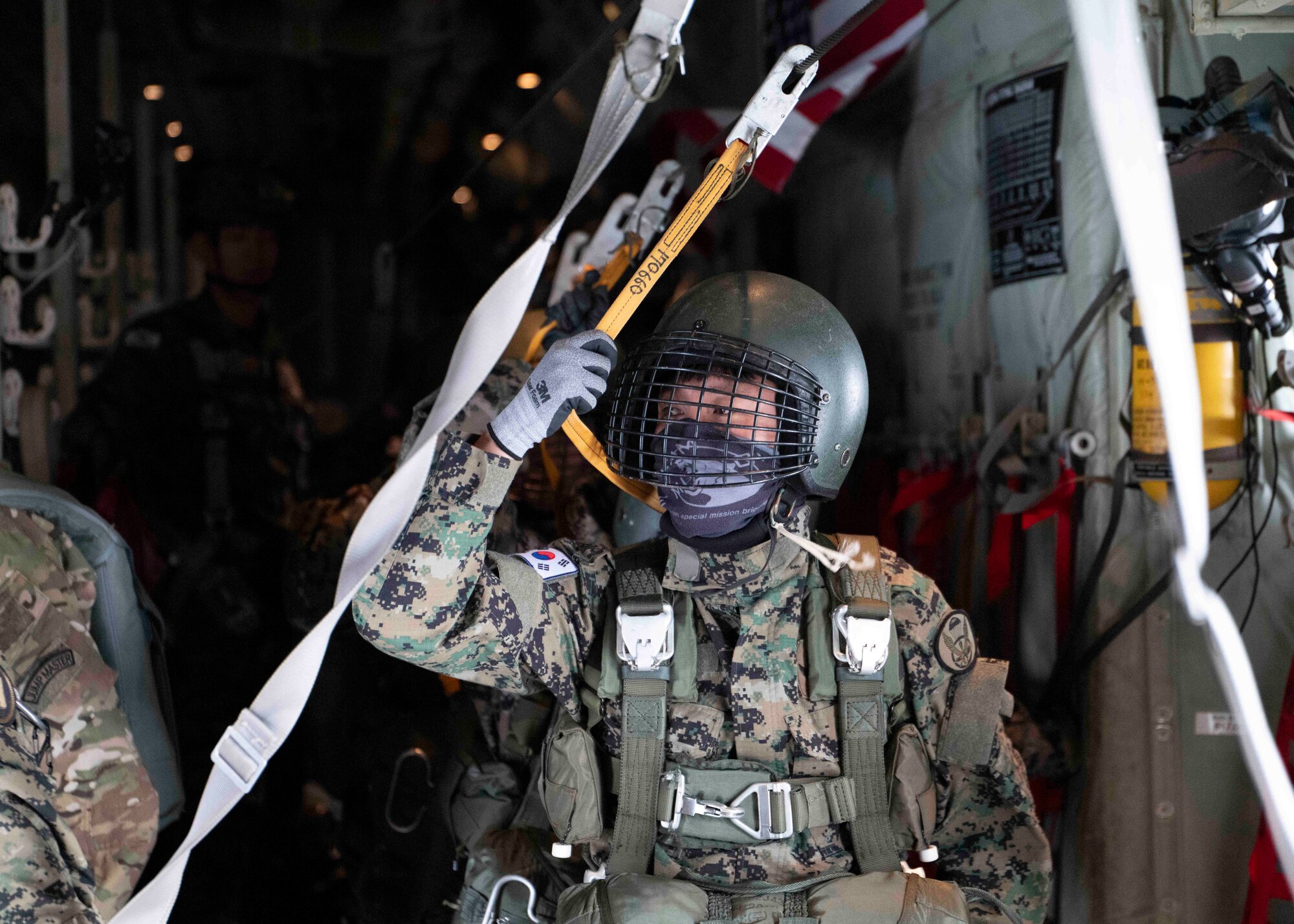 A uniformed man wearing a helmet, gloves, and parachute rigging stands in line inside of a C-130.