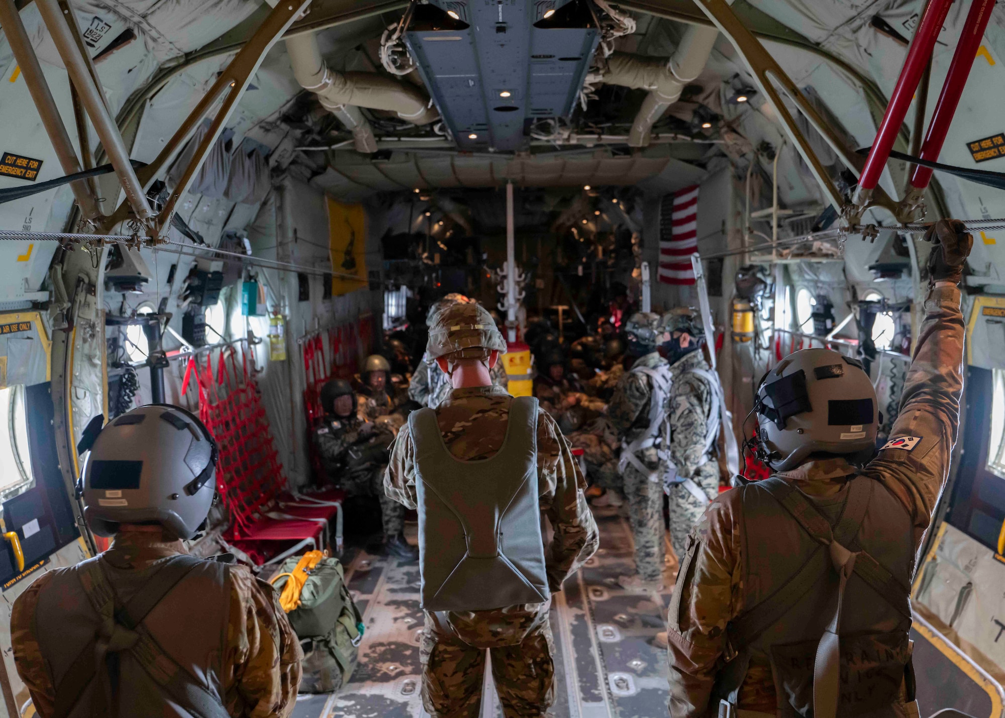 Three men wearing flight suits and parachute rigging face away from the camera and toward two rows of uniformed men sitting facing each other in the cargo hold of a C-130J.