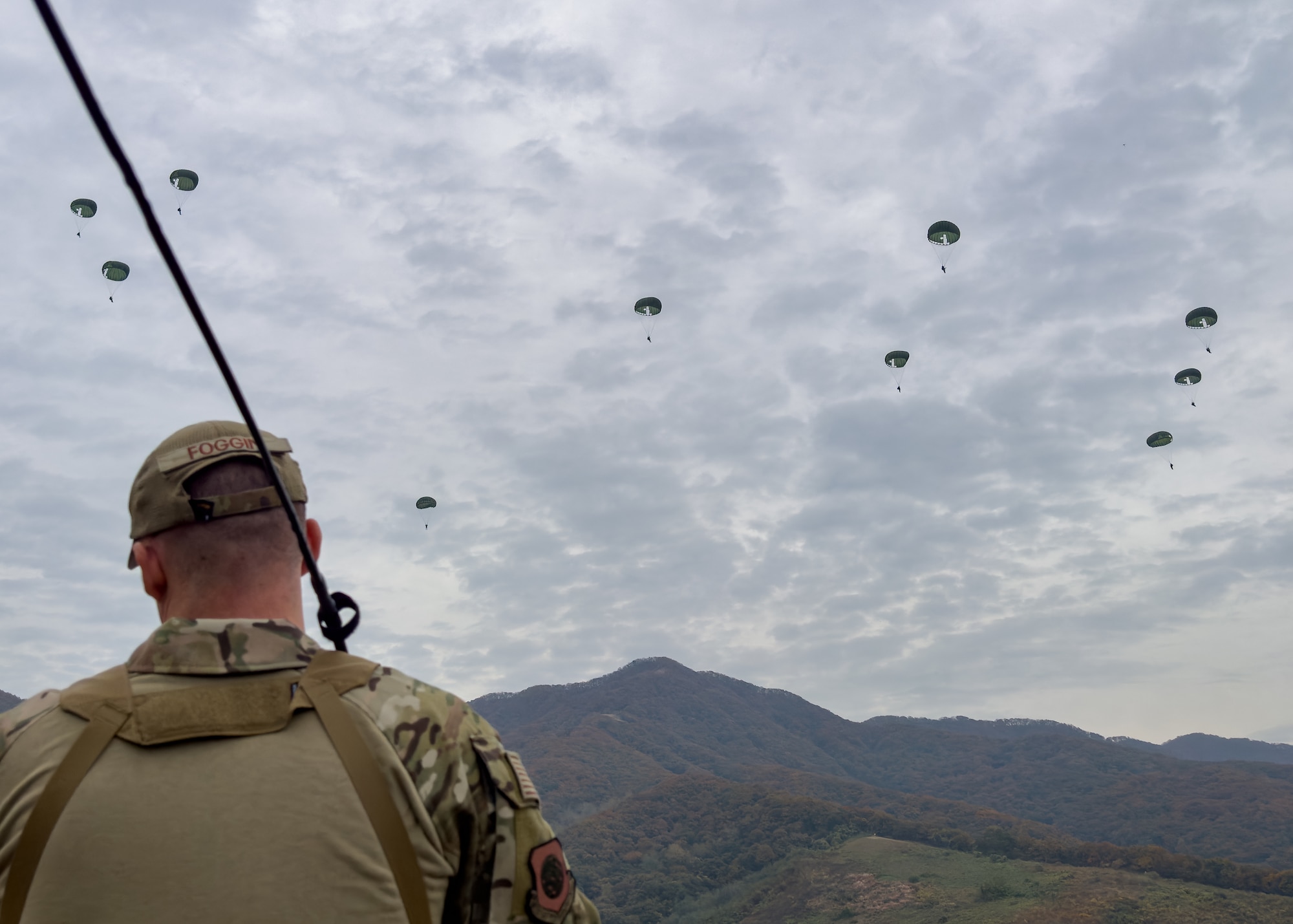 A uniformed man looks out over a mountain with 10 parachute jumpers in the gray sky.