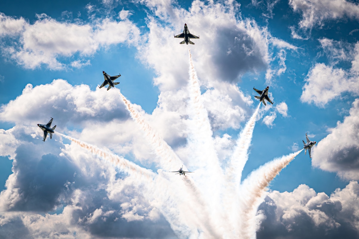 Six military fighter aircraft soar in sync against a cloudy sky leaving streaks of smoke in their wake.