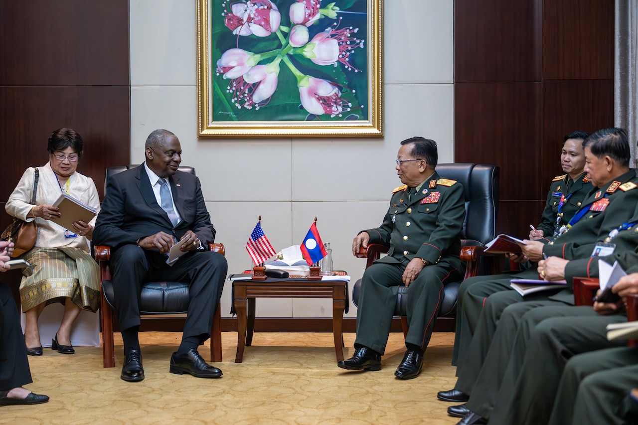 A man wearing business attire sits next to a man wearing a foreign military uniform.