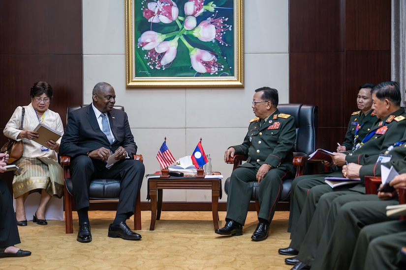 A man wearing business attire sits next to a man wearing a foreign military uniform.