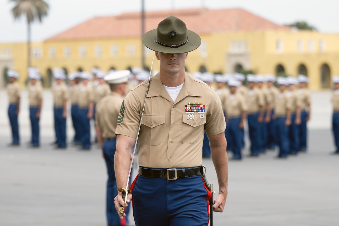 A Marine walks with a sword over his shoulder in front of a group of Marines at attention.