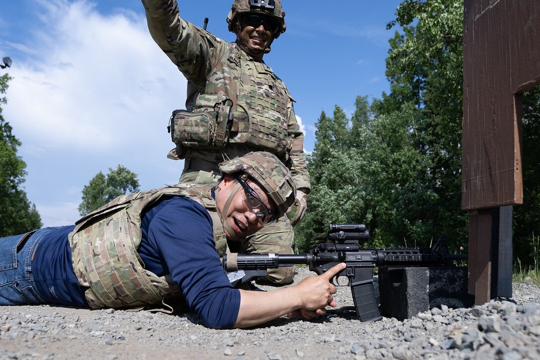 A civilian wearing combat hear and holding a weapon lays on the ground while a service members instructs from above.