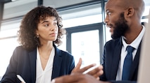 A black woman and a black man dressed in suits, sitting at a meeting together discussing work