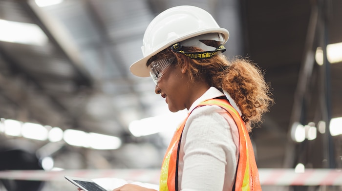 A black woman checking her tablet in a logistics department, wearing a hard hat and orange vest