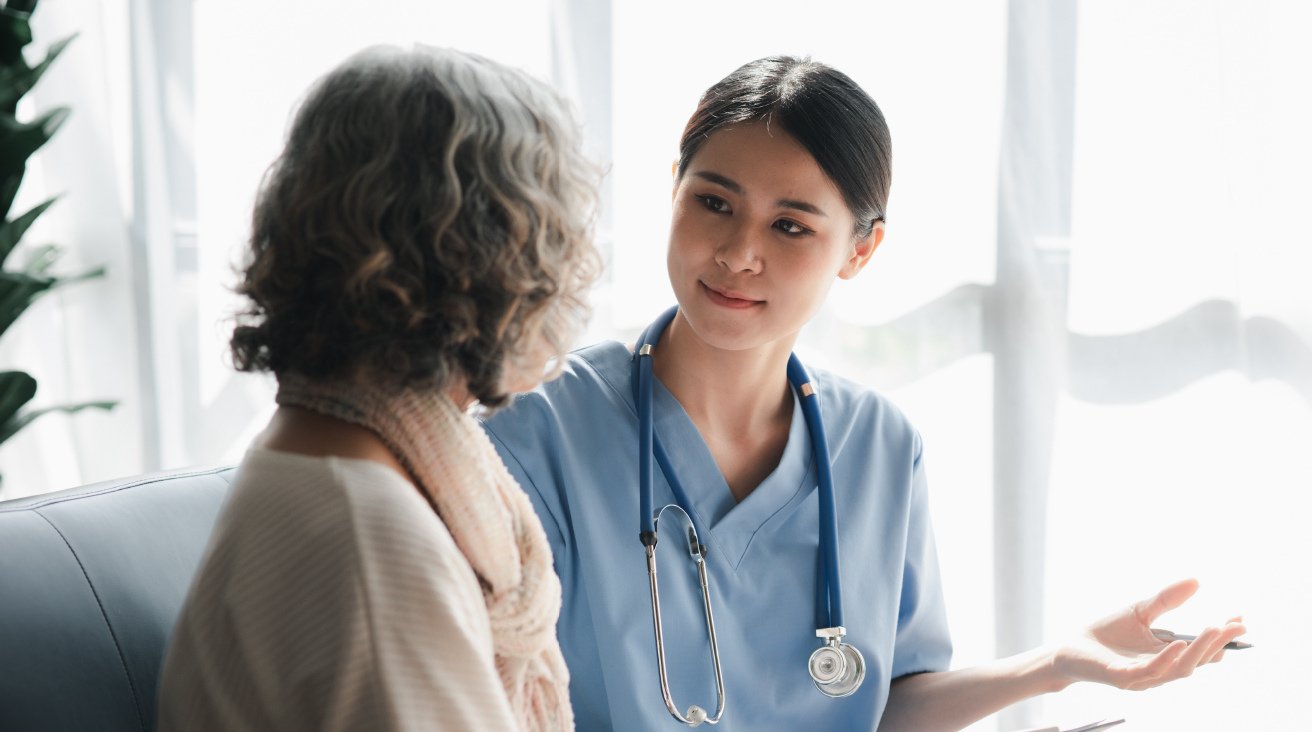 A woman doctor wearing scrubs and a stethoscope explaining information to a gray-haired woman who is sitting next to her.