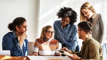 A group of diverse college students sitting and standing around a desk discussing a project