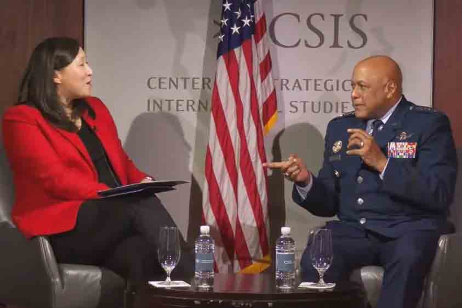 A woman and a man speak while sitting at a table on a stage.