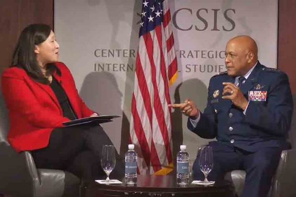 A woman and a man speak while sitting at a table on a stage.