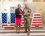 man and woman holding a plaque and smiling in front of a USA flag