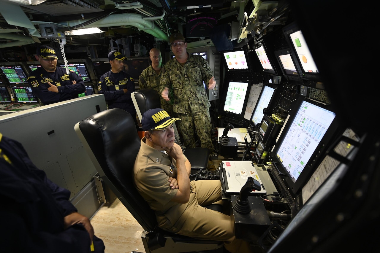 A man sits at the helm of a room filled with monitors as others around him talk.