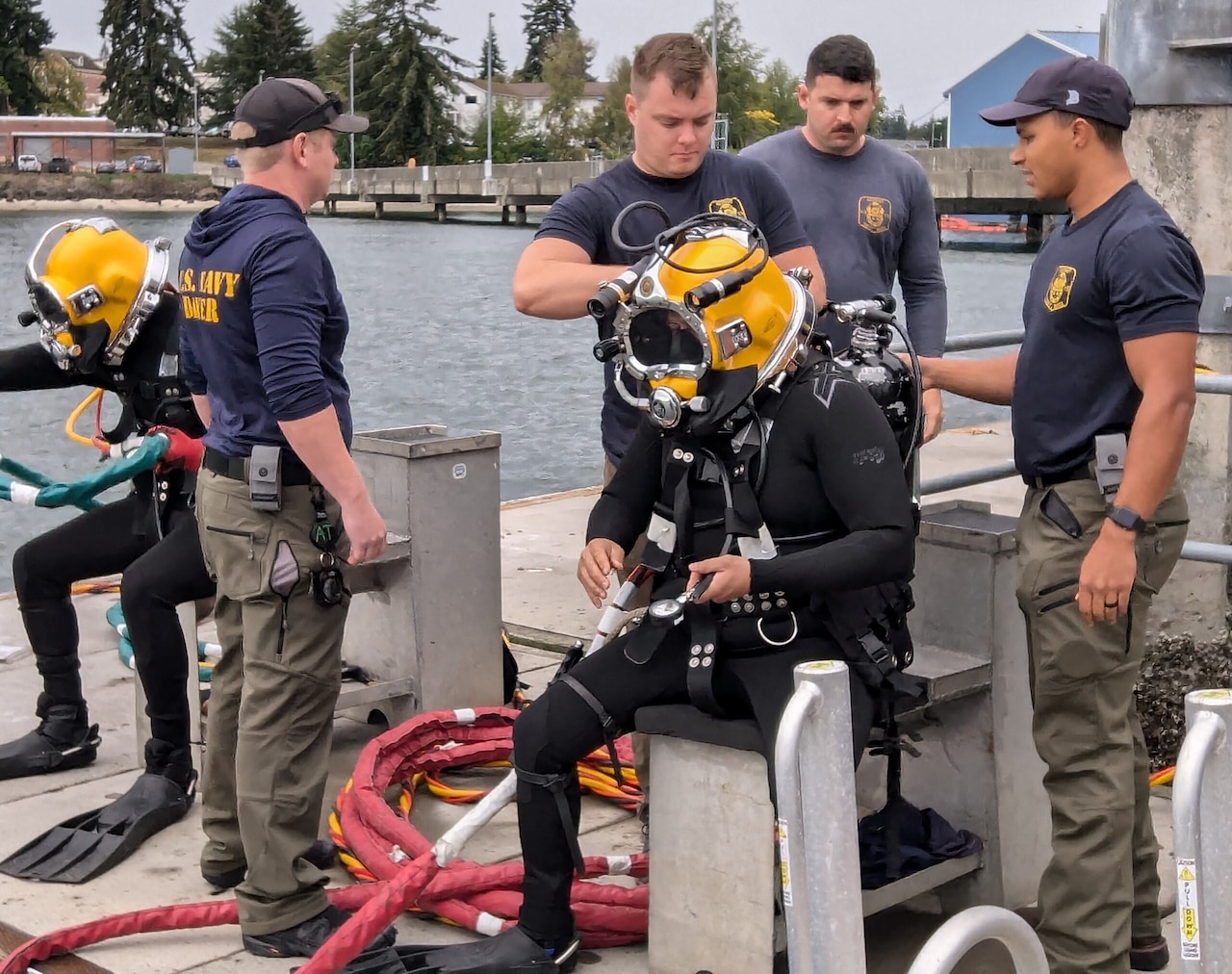 Two men in wetsuits and dive helmets sit while others help them with their gear.