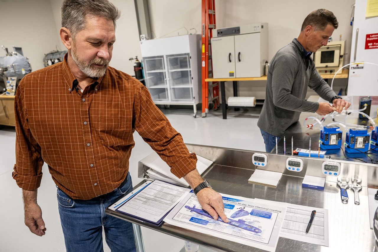 A man points to a printout while another man attaches tubes to a device in the background.