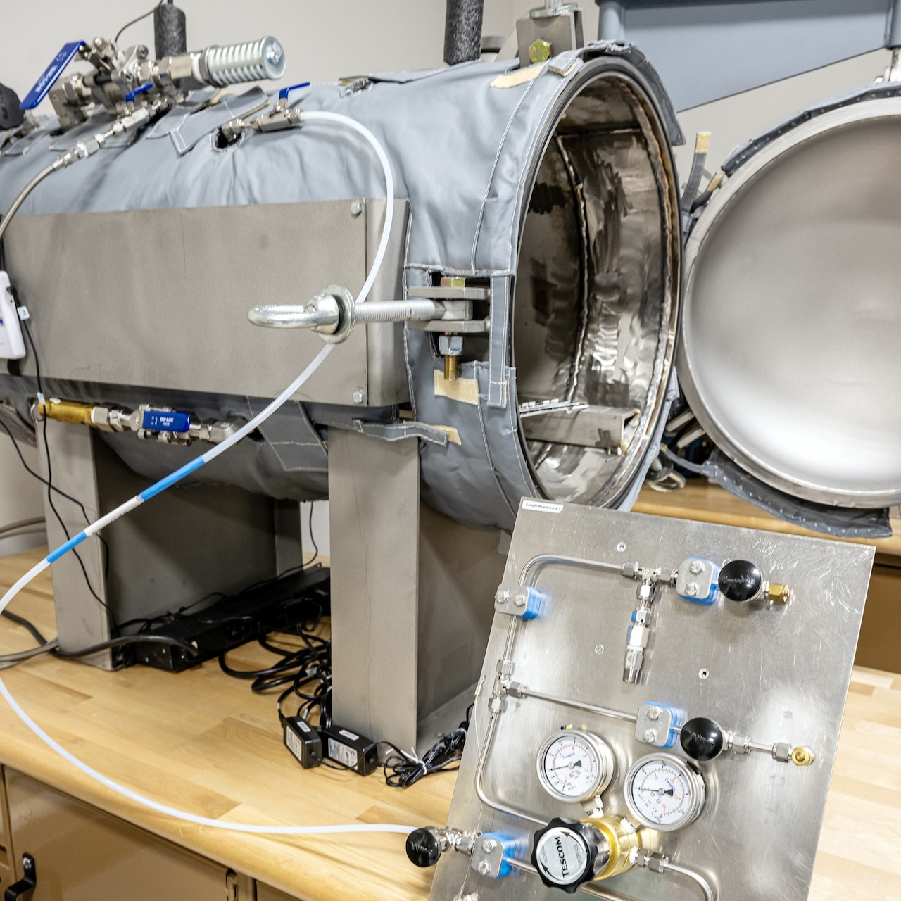 A cylindrical metal chamber with its door open is attached to tubes and gauges.