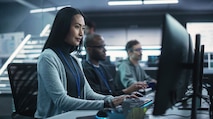 An Asian woman working next to two men all sitting at desks with computers, analyzing data