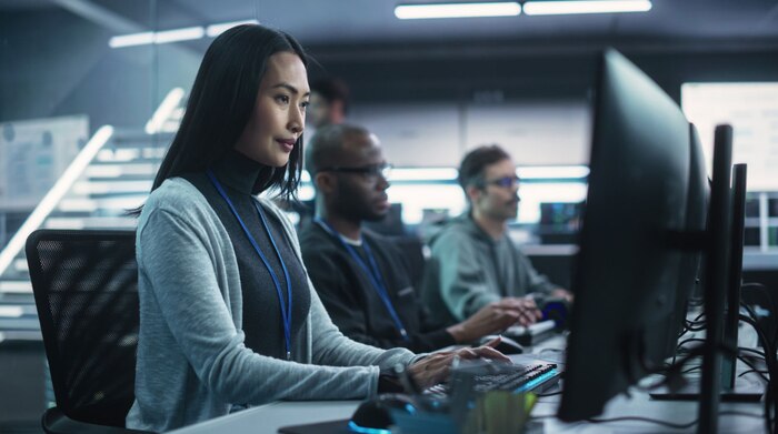 An Asian woman working next to two men all sitting at desks with computers, analyzing data