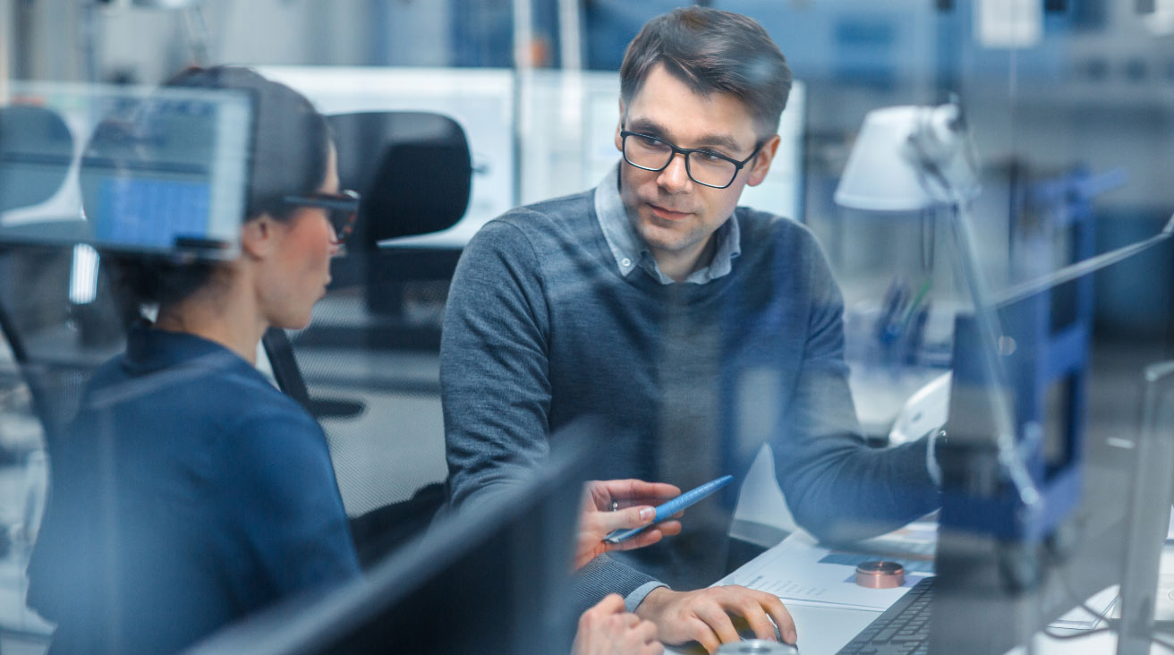 A man and woman engineer having a conversation while sitting at a desk with computers in front of them.