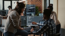 An NSA employee sitting on a desk speaking to two summer interns about data on their computer screen