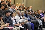 A group of professionally dressed people sitting in a large room while someone sits with a microphone in their hands.