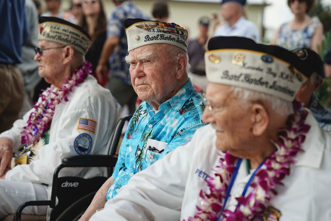 Three veterans wearing caps and leis sit at an outdoor ceremony.