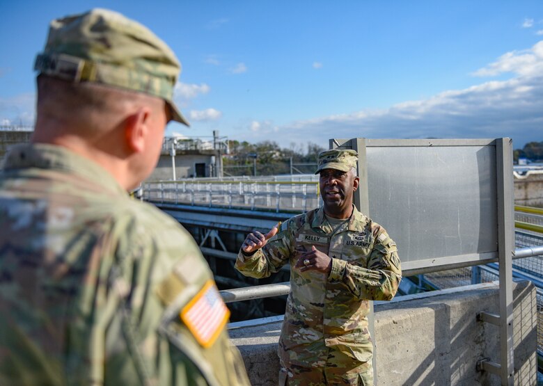 Two men in military fatigues converse in front of a bridge.