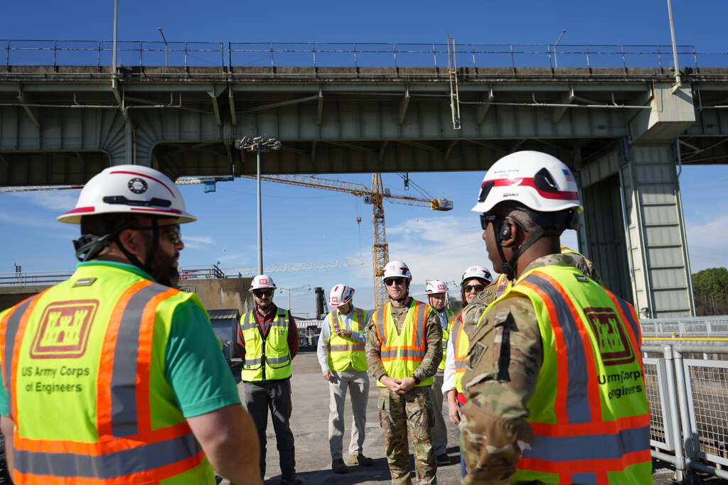People in safety vests and hardhats, including both civilians and U.S. Army servicemembers, talk underneath a bridge.