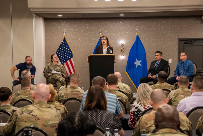 Air Force civilian on a stage with one Airmen and three other civilians addressing an audience from a podium during a conference.