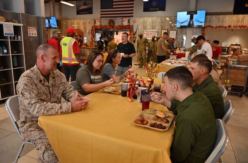 A uniformed service member sits at a table while several others eat.