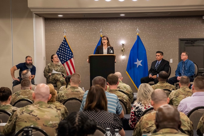 Air Force civilian on a stage with one Airmen and three other civilians addressing an audience from a podium during a conference.