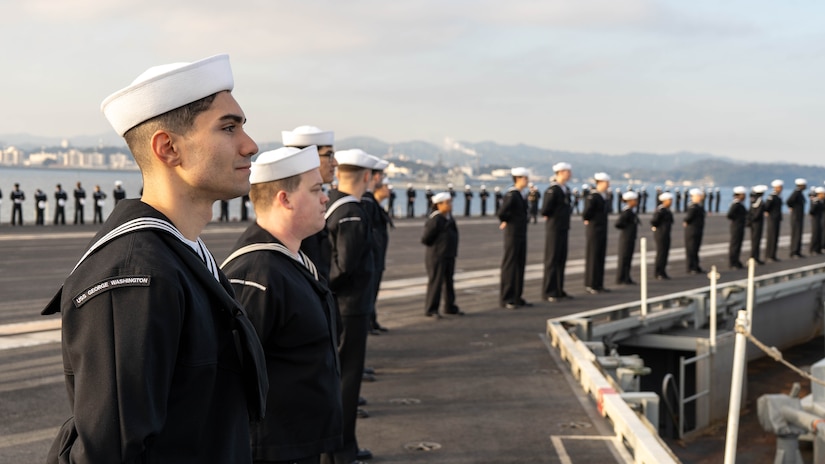 Sailors man the rails of an air craft carrier during a cloudy day.