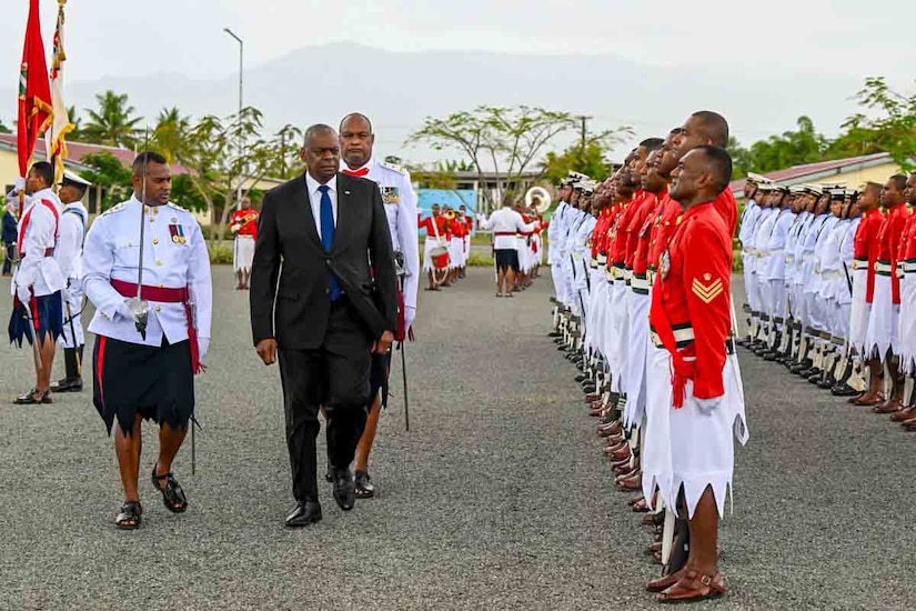 A man in business attire walks next to a formation of foreign service members in uniform.