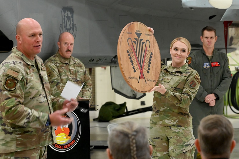 An airman smiles and holds a piece of artwork while other airmen stand nearby.