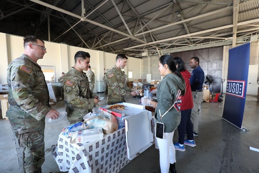 Soldiers and civilians line up beside a long table with boxes of pizza on it.